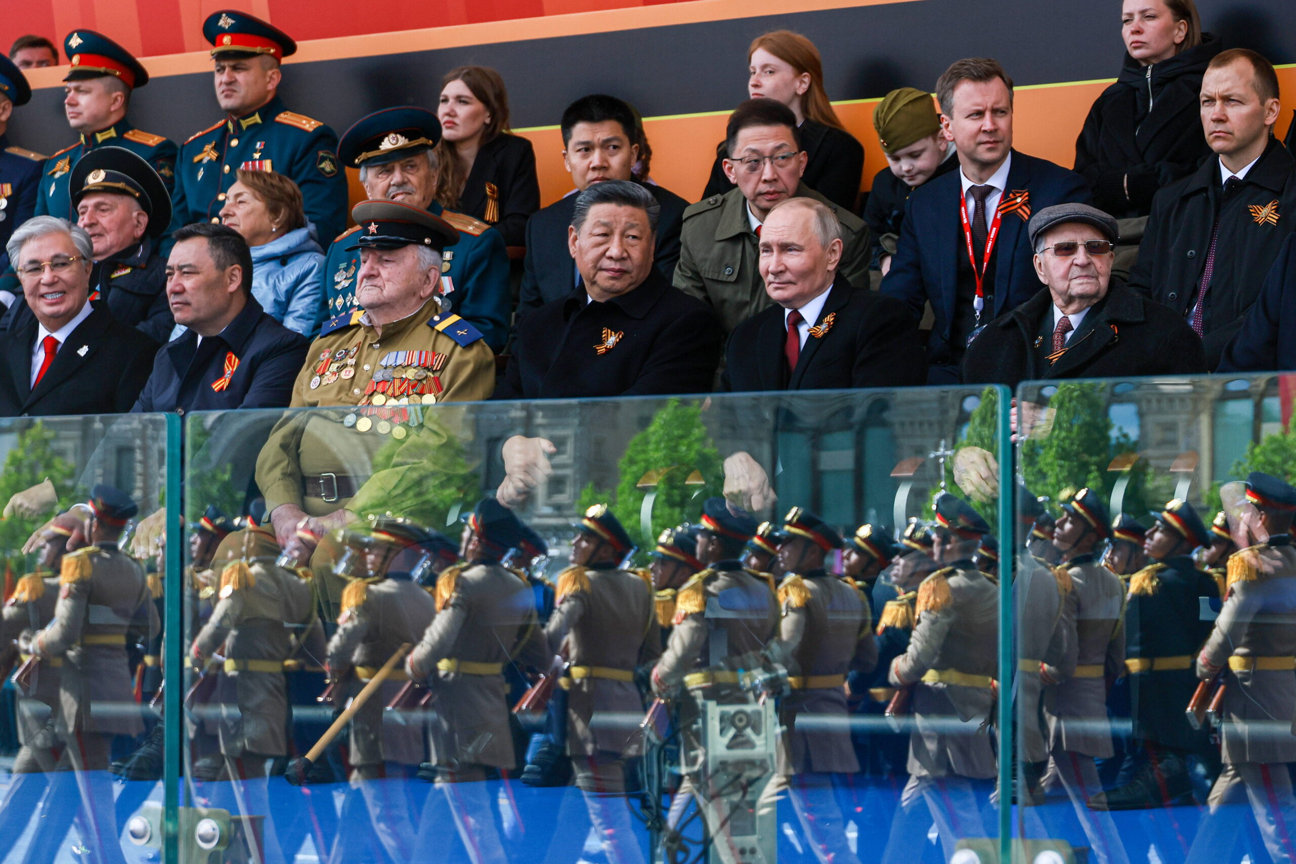 Xi Jinping et Vladimir Poutine côte à côte au premier rang de la tribune, encadrés de vétérans décorés et d’autres officiels, tandis que le défilé se reflète dans la vitre de protection. Photo : Vladimir Smirnov / ZUMA