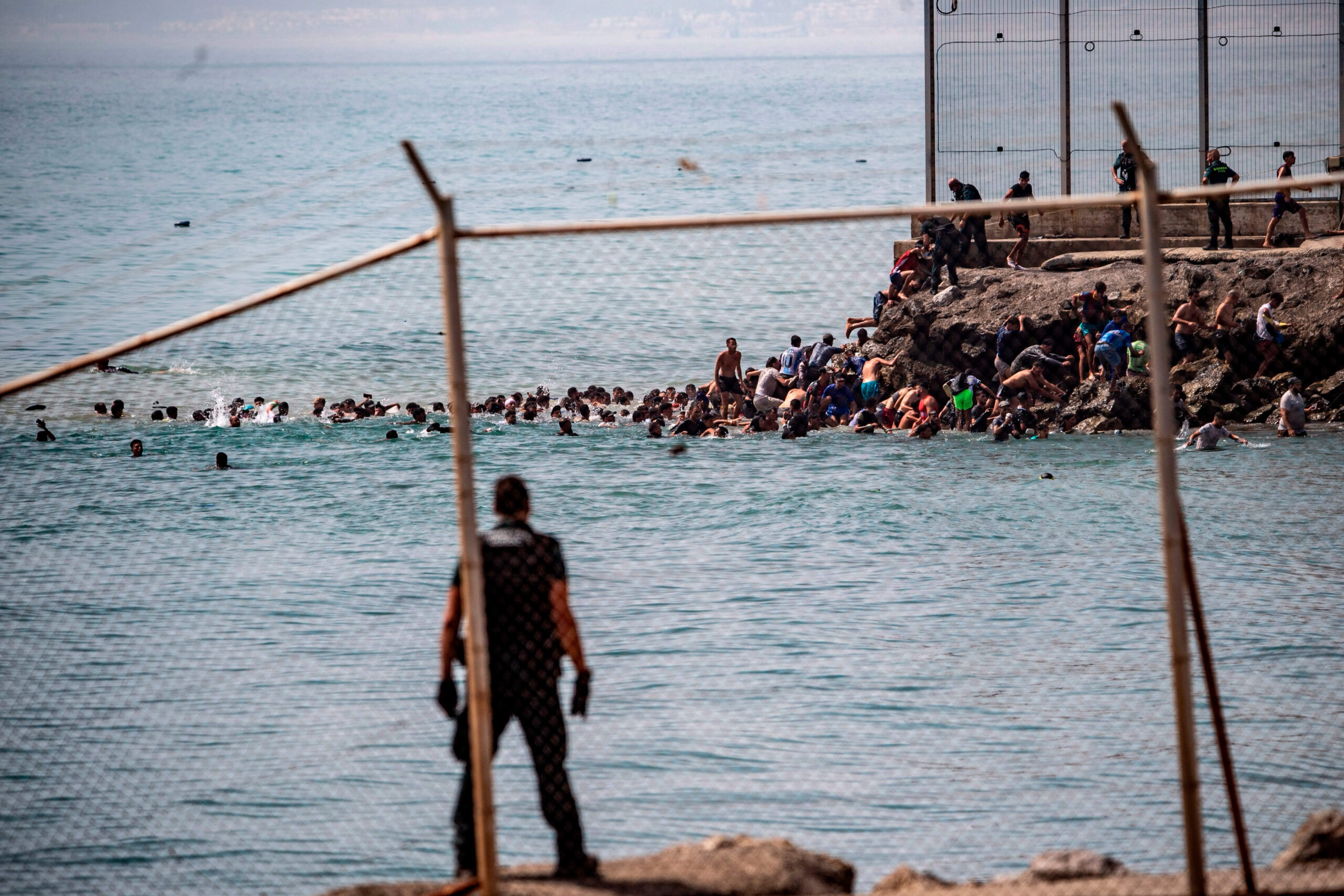 Vue à travers le grillage frontalier : sous le regard des agents, des dizaines de nageurs contournent l'extrémité de la clôture pour prendre pied sur les rochers de Ceuta, jeudi 30 juillet 2026. © AP Photo/Antonio Sempere/SIPA