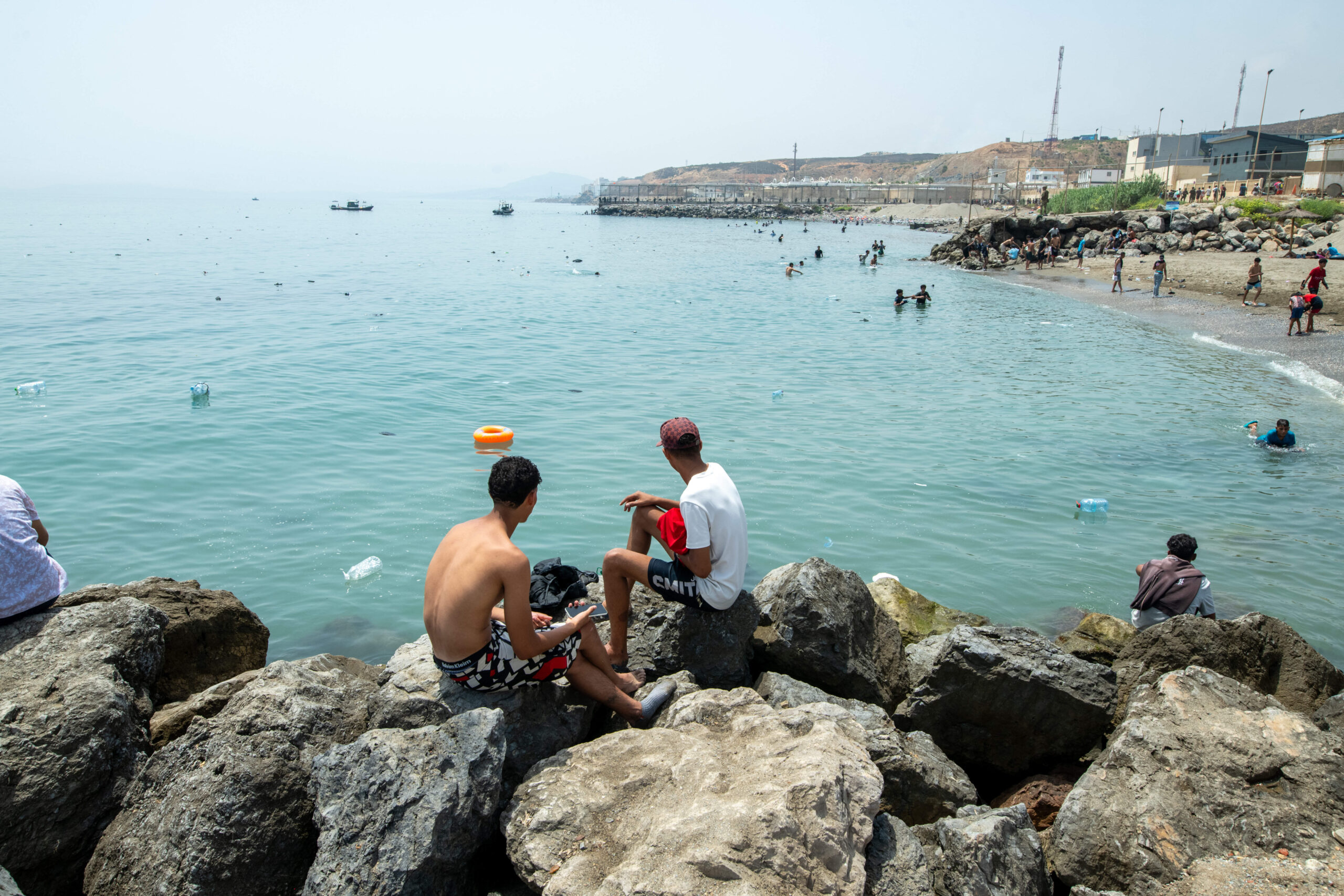 Depuis les rochers, de jeunes marocains contemplent une baie qui ressemble à n’importe quelle plage du détroit, à ceci près que le grillage frontalier, au fond, coupe le littoral en deux et plonge dans la mer. Photo : Juan Carlos Lucas