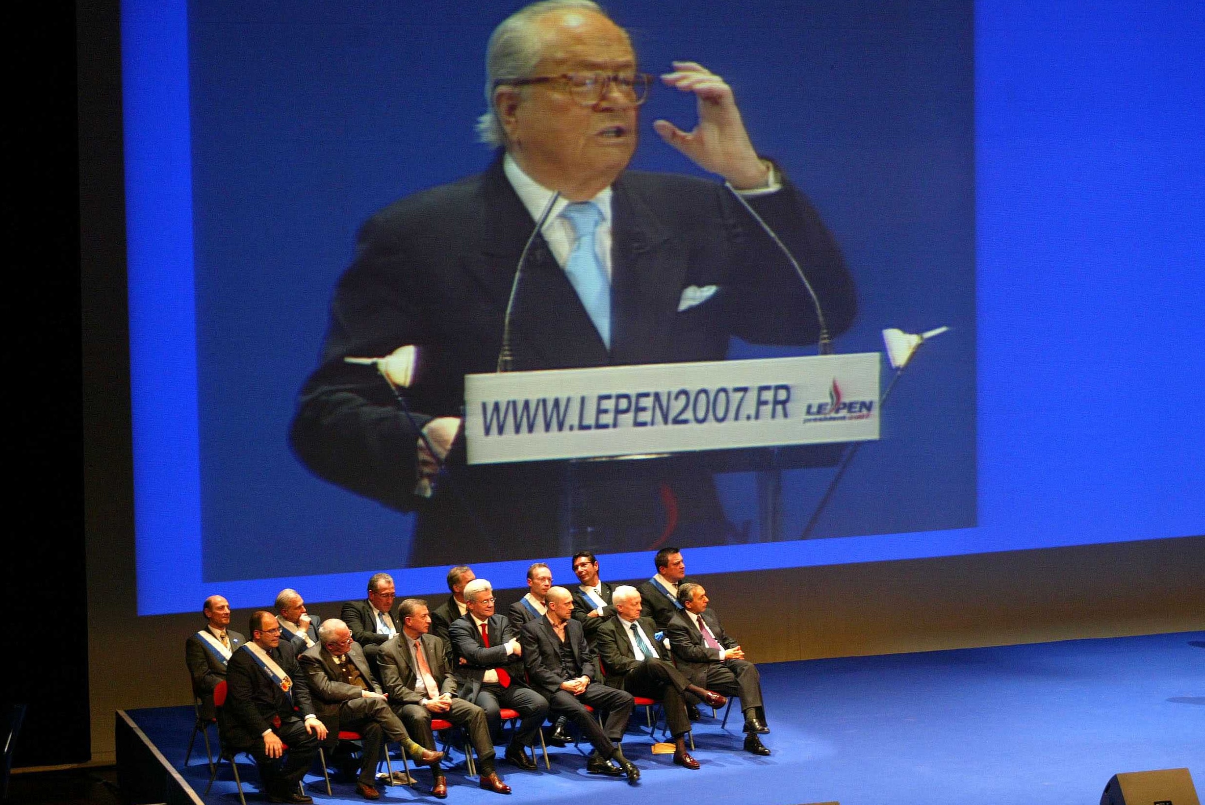 Jean-Marie Le Pen delivers a speech during this meeting, transcribed on a large screen. Alain Soral is on stage.